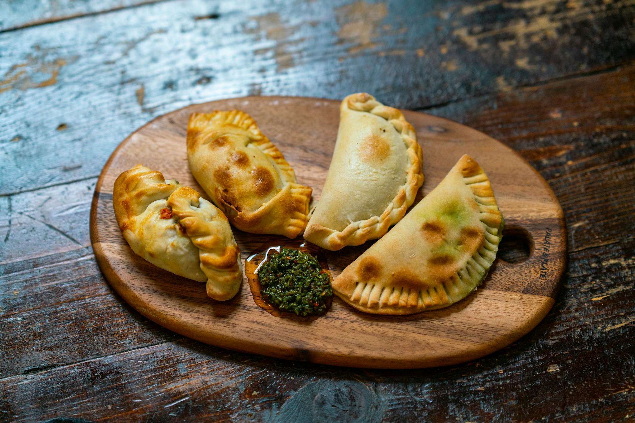 Appetizing baked empanadas served with chimichurri sauce on a wooden board. Perfect for food lovers.